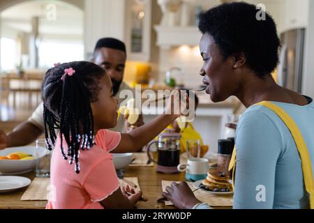 Ein afroamerikanisches Mädchen füttert die Mutter mit Pfannkuchen, während es mit der Familie am Esstisch frühstückt Stockfoto