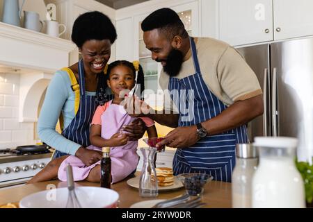 Fröhlicher afroamerikanischer Mann, der seiner Tochter Obst fütterte, von der Mutter auf dem Küchentisch gehalten Stockfoto