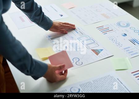 Nahaufnahme einer jungen Büroleiterin mit Notizpapier in der Hand, die sich über den Schreibtisch beugt, mit Finanzpapieren mit verschiedenen Diagrammen und Diagrammen Stockfoto