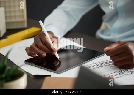 Konzentrieren Sie sich auf die Hand eines jungen afroamerikanischen Architekten, der eine Skizze eines neuen Gebäudes mit Eingabestift auf dem Tablet-Bildschirm zeichnet, während er an seinem Arbeitsplatz sitzt Stockfoto