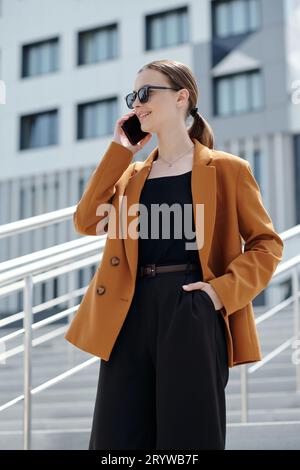 Junge lächelnde Geschäftsfrau in braunem Blazer und schwarzer Weste und Hose, die mit jemandem auf dem Smartphone spricht, während sie auf der Treppe steht Stockfoto