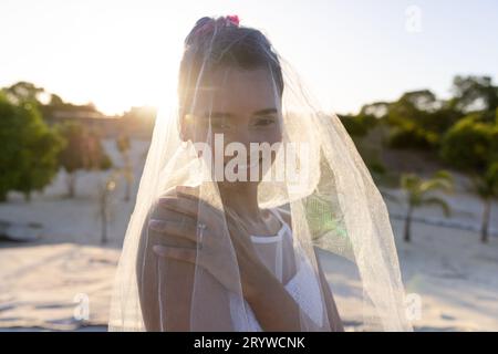 Porträt einer schönen zweirassigen jungen Braut, die einen Schleier trägt und am Strand vor klarem Himmel steht Stockfoto