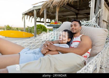 Lächelndes, birassisches junges Paar, das wegblickt und sich in der Hängematte am Strand entspannt, Kopierraum Stockfoto