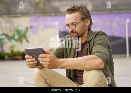 Mann mittleren Alters, der im Freien sitzt und von einem Tablet-PC aus studiert. Mit einem fokussierten Ausdruck nutzt er die digitale Entwicklung Stockfoto