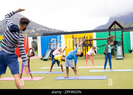 Verschiedene männliche Lehrer und Grundschulkinder, die Yoga auf dem Schulhof praktizieren Stockfoto