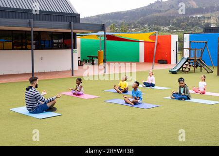 Diverse männliche Lehrer und Grundschulkinder, die Meditation auf dem Schulhof praktizieren Stockfoto