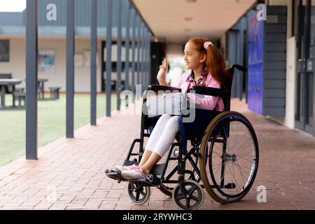 Glückliches birassisches Schulmädchen im Rollstuhl, winkend im Schulflur Stockfoto