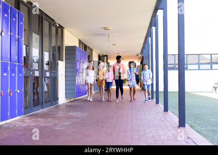 Glückliche, vielfältige Lehrerinnen und Schulkinder, die zusammen im Schulkorridor laufen Stockfoto