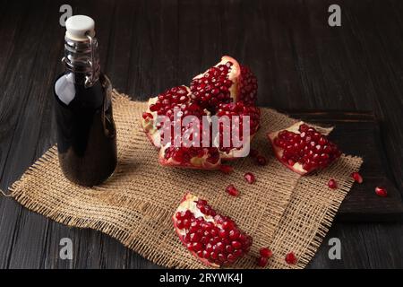 Reifer Granatapfelbruch, verstreute große Körner, frisch gepresster Granatapfelsaft in einer transparenten Flasche auf Holzhintergrund. Stockfoto