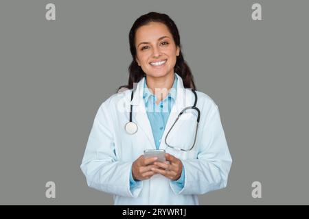 Ärztin, die ein Smartphone mit medizinischer Anwendung hält, auf grauem Studiohintergrund steht und in die Kamera lächelt Stockfoto