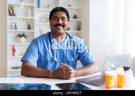 Lächelnder Indischer Männlicher Arzt In Blauer Uniform, Der Am Schreibtisch Mit Laptop Sitzt Stockfoto