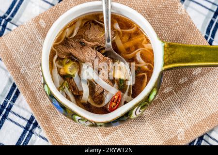 Traditionelle vietnamesische Suppe Pho Bo mit Rindfleischstück, Reisnudeln und Kräutern in Schüssel auf Handtuch auf kariertem Tischtuch, Nahaufnahme. Stockfoto