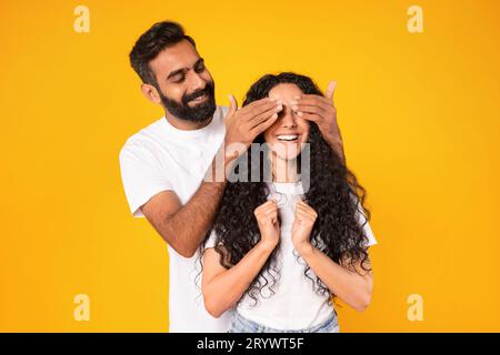 Mann aus dem Nahen Osten, der die Augen der Frau bedeckt, überrascht sie, gelber Hintergrund Stockfoto