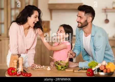 Fröhliche Tochter füttert ihre glückliche Mutter mit kleinen Kirschtomaten, während sie gemeinsam mit den Eltern Salat zubereitete Stockfoto