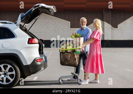 Liebevolles älteres Paar, das neben dem Auto mit Lebensmittelpaketen steht Stockfoto