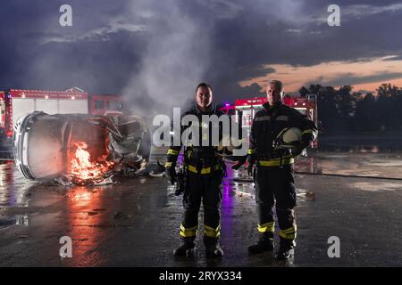 Mutiges Feuerwehrteam geht zur Kamera. Im Hintergrund kämpfen Sanitäter und Feuerwehrleute Rettungsdienst Feuer bei Autounfall, INSU Stockfoto