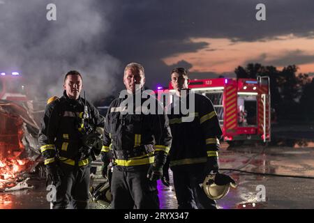 Mutiges Feuerwehrteam geht zur Kamera. Im Hintergrund kämpfen Sanitäter und Feuerwehrleute Rettungsdienst Feuer bei Autounfall, INSU Stockfoto