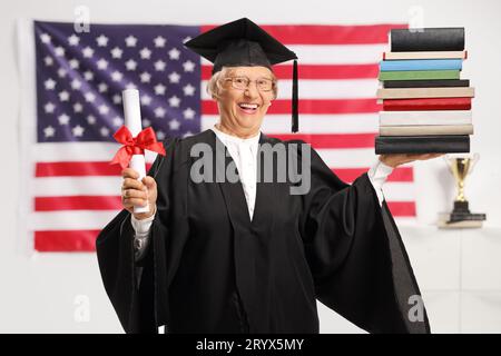 Seniorin in einem Abschlusskleid mit einem Diplom und einem Stapel Bücher mit der Flagge der USA im Hintergrund Stockfoto