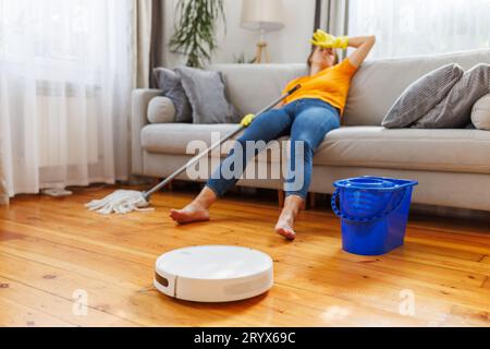Ein Roboter-Staubsauger reinigt ein Zimmer, während eine müde Frau sich auf dem Sofa entspannt Stockfoto