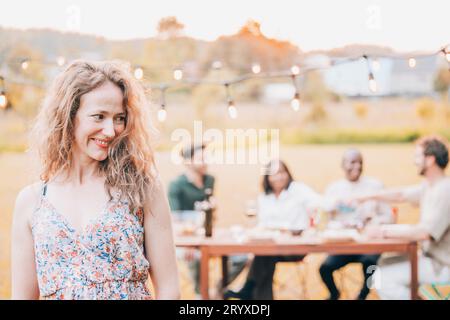 Schöne junge Frau mit lockigen blonden Haaren in einem Sommerkleid mit einer Gruppe von Freunden im Hintergrund, die einen Garten im Freien haben Stockfoto