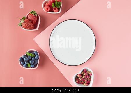 Leerer Teller und kleine Teller in Form von Herzen mit frischen Erdbeeren, Himbeeren, Heidelbeeren auf rosafarbenem Hintergrund. Obere V Stockfoto