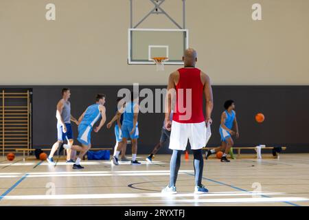 Verschiedene männliche Basketballspieler, die blaue Sportkleidung tragen und mit Trainer im Fitnessstudio Basketball spielen Stockfoto