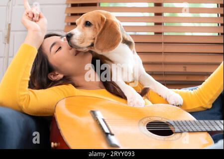 An einem Wochenende spielen Eine junge Asiatin und ihr Beagle-Welpe Gitarre und singen fröhlich zusammen im Wohnzimmer Stockfoto