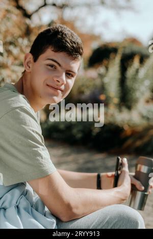 Ein Teenager sitzt auf Einer Bank im Autumn Park und trinkt Kaffee aus Einer Thermo-Tasse und schaut in Ein Telefon. Porträt Von Gutaussehendem Beifall Stockfoto