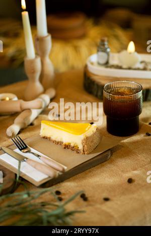 Neben einem Stück Orangenkäsekuchen sitzt auf einem Holzbrett auf einer Sackleinen-Tischdecke ein eisiger Americano im Glas, der durch das warme Schein von b ergänzt wird Stockfoto