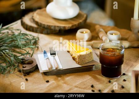 Neben einem Stück Orangenkäsekuchen sitzt auf einem Holzbrett auf einer Sackleinen-Tischdecke ein eisiger Americano im Glas, der durch das warme Schein von b ergänzt wird Stockfoto