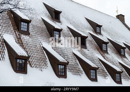 Schnee fällt auf die Dächer der Stadt. Schneebedecktes braunes Metallziegeldach des europäischen Hauses mit Fenstern. Überdacht auf dem Dach Stockfoto