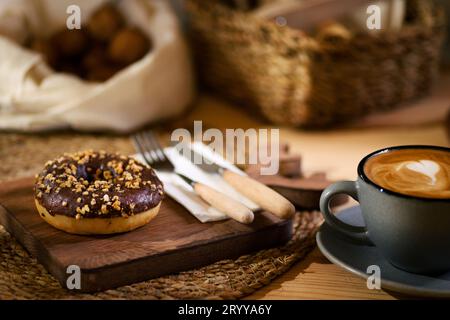 Ein Donut auf einem Holzbrett, neben einem Latte in einer grauen Tasse. Weiße Serviette mit Holzbesteck und Schalenfrüchten in weißem Tuch im Hintergrund. Stockfoto
