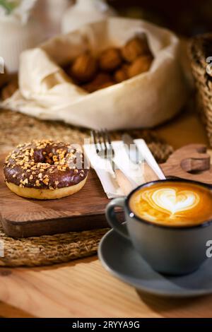 Ein Donut auf einem Holzbrett, neben einem Latte in einer grauen Tasse. Weiße Serviette mit Holzbesteck und Schalenfrüchten in weißem Tuch im Hintergrund. Stockfoto