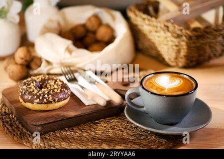 Ein Donut auf einem Holzbrett, neben einem Latte in einer grauen Tasse. Weiße Serviette mit Holzbesteck und Schalenfrüchten in weißem Tuch im Hintergrund. Stockfoto