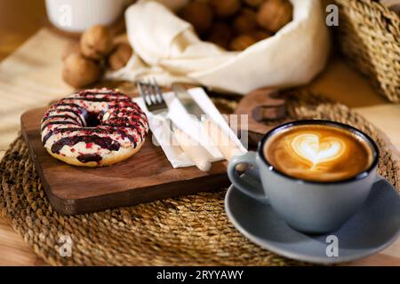 Ein Donut auf einem Holzbrett, neben einem Latte in einer grauen Tasse. Weiße Serviette mit Holzbesteck und Schalenfrüchten in weißem Tuch im Hintergrund. Stockfoto