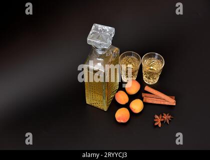 Aprikosenlikör mit Gewürzen, starker hausgemachter Alkohol in einer Kristallkaraffe und zwei Gläser auf schwarzem Hintergrund, neben Anis und Zimtstangen. C Stockfoto