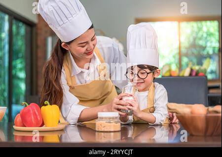 Asiatische Mutter hilft süßen kleinen Jungen, der Milch im Glas in der Küche zu Hause in Koch-Uniform trinkt. Die Menschen leben und sind glücklich Stockfoto