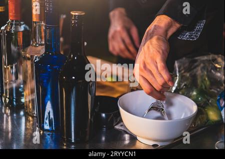 Nahaufnahme der alkoholischen Flaschen mit Barmixer Hand, die frischen Limettenlimonade Cocktail in Trinkweinglas mit Eis in der Nacht zubereiten Stockfoto