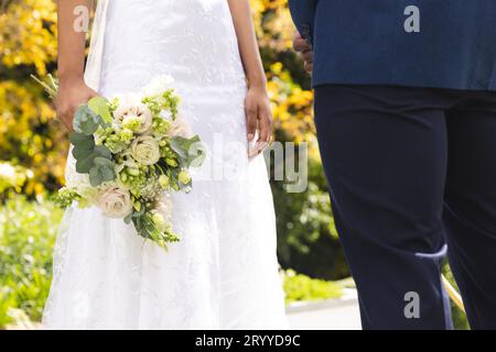 Mittelteil der afroamerikanischen Braut mit Blumenstrauß und Bräutigam im sonnigen Garten Stockfoto