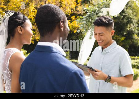 Glücklicher, vielseitiger Beamter mit Tablet, mit Braut und Bräutigam bei der Hochzeitszeremonie im sonnigen Garten Stockfoto