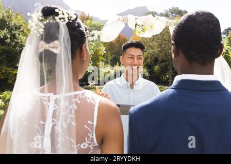 Glücklicher, vielseitiger Beamter mit Tablet, mit Braut und Bräutigam bei der Hochzeitszeremonie im sonnigen Garten Stockfoto