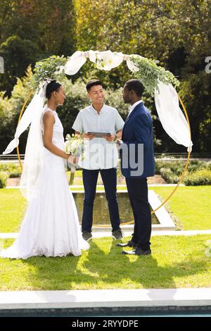 Ein vielseitiger männlicher Beamter, Braut und Bräutigam bei der Hochzeitszeremonie im sonnigen Garten Stockfoto