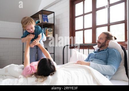 Kleiner Junge, der mit Mom flog, benutzte ihre Beine, um ihm zu helfen, hoch vom Boden seines Bettes im Schlafzimmer zu fliegen. Stockfoto