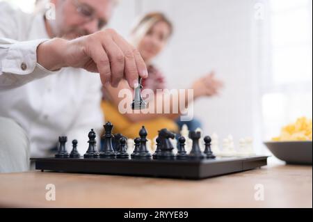 Im Wohnzimmer des Hauses sitzt ein älteres Paar und entspannt sich. Um gemeinsam mit einem Schachbrett mit einem d zu spielen Stockfoto