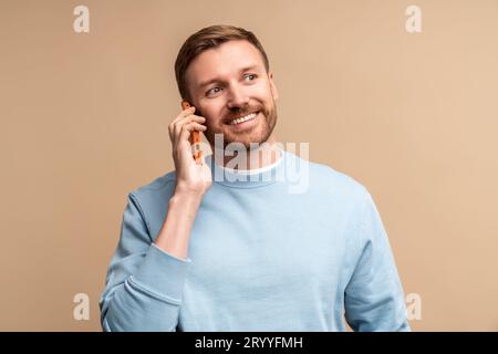 Glücklicher lächelnder Mann mittleren Alters, der auf beigefarbenem Hintergrund telefoniert und beiseite schaut. Stockfoto