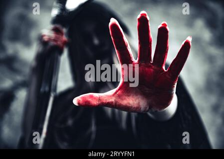 Hexe zeigt blutige Hand mit Sensenmann. Weiblicher Dämonenenengel in schwarzer Kleidung und Kapuze auf Grunge-Wand-Hintergrund. Halloween Tag und Stockfoto