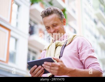 Glücklicher Mann, der die neuesten Nachrichten von einem Tablet-PC liest, während er draußen auf der Stadtstraße ist. Der Tablet-PC in seinen Händen ermöglicht es ihm Stockfoto