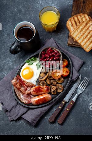 Englisches Frühstück mit Spiegeleiern, Würstchen, Speck, Pudding, Bohnen, Toast und Kaffee, dunkler Hintergrund Stockfoto
