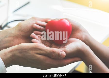 Arzthände halten und geben dem Patienten kleine Kinder ein rotes Massageherz, um sich von der Krankheit zu erholen. Krankenhaus und Gesundheitswesen Stockfoto