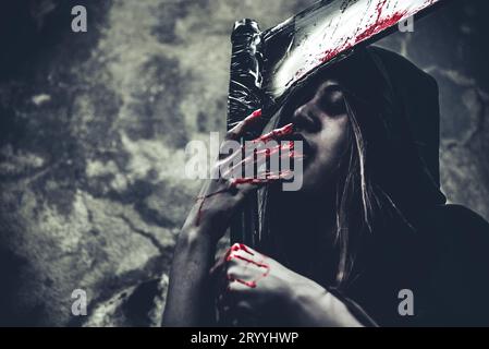 Hexe leckt Blut an den Fingern. Weiblicher Dämonenenengel in schwarzer Kleidung und Kapuze auf Grunge-Wand-Hintergrund. Halloween-Tag und Myster Stockfoto